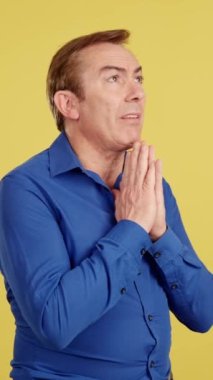 Mature man praying with folded hands in studio with yellow background