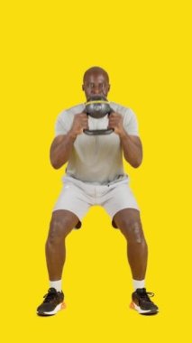 Stüdyoda Afrikalı bir adamın sarı arkaplanlı videosu var. Kettlebell 'li globet çöreği yapıyor.