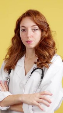 Kameraya gülümseyen kızıl saçlı kadın doktor. Stüdyoda kollarını kavuşturmuş, sarı arka planda.