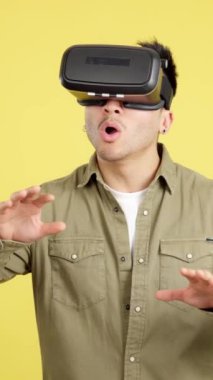 Sanal gerçeklik gözlüklü mutlu Çinli adam sarı arka planlı stüdyoda sürpriz yapıyor.