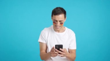 Yüzünde Alman bayrağı olan, cep telefonuna bakıp gülümseyen ve krom tuşlu ekranla kameraya gösteren bir Alman 'ın krom arkaplan görüntüsü..