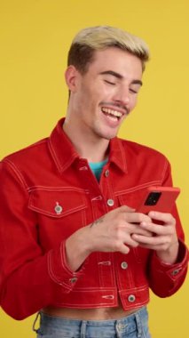 Stüdyoda sarı arka planda cep telefonunu kullanırken gülümseyen mutlu gay adam.