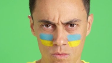 Stüdyoda, yüzünde Ukrayna bayrağı olan ve kameraya bakan ciddi bir adamın krom görüntüsü var.