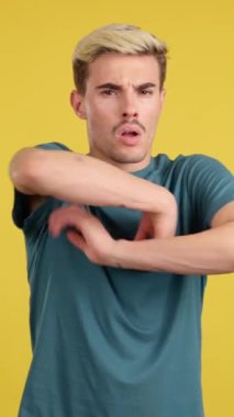 Stüdyoda sarı arka planda kızgın bir ifadeyle kollarını birleştiren eşcinsel adam.