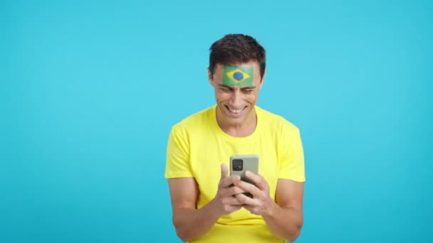 ブラジルの旗が顔に塗られたブラジル人のクロマの背景のスタジオビデオ 彼の携帯電話を笑顔に見て クロマキーのスクリーンでカメラにそれを示す — ストック動画