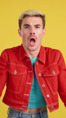 Stüdyoda sarı arka plandaki kameraya bakıp el kol hareketi yapan eşcinsel bir adam.