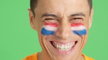 Stüdyoda, yüzünde Hollanda bayrağı olan ve kameraya gülümseyen bir adamın krom görüntüsü var.
