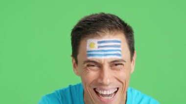 Stüdyoda, yüzünde uruguay bayrağı olan ve kameraya gülümseyen bir adamın krom görüntüsü var.