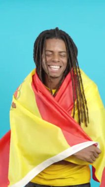 Stüdyoda mavi arka planda İspanyol bayrağıyla sarılan rastalı mutlu Latin adam.