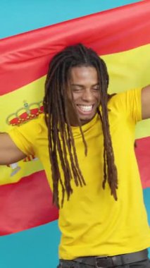 Rastalı mutlu Latin adam gülümsüyor ve mavi arka planlı bir İspanyol bayrağını göndere çekiyor.