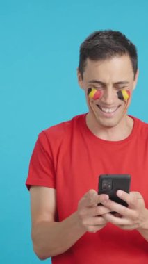 Yüzünde Belçika bayrağı olan Belçikalı bir destekçinin krom arkaplanlı stüdyo videosu, cep telefonuna bakıp gülümsüyor ve krom tuşlu ekranla kameraya gösteriyor.