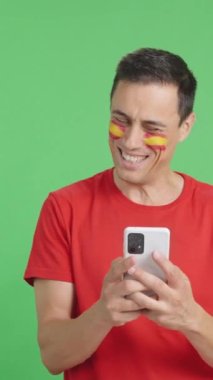 Yüzünde İspanya bayrağı olan, cep telefonuna bakıp gülümseyen ve ekranı krom tuşlu gösteren İspanyol bir adamın krom arkaplanlı stüdyo videosu..