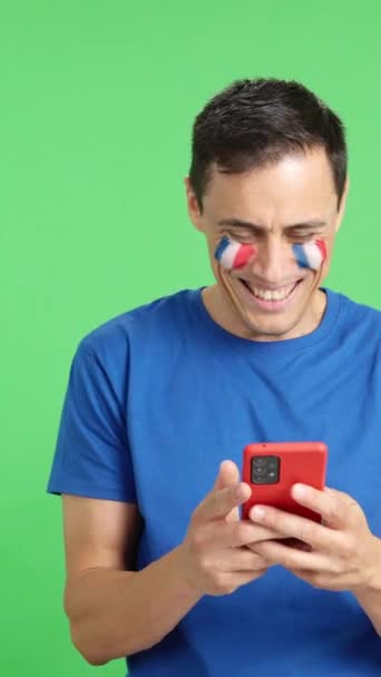 フランスの旗を持つフランスの男のクロマの背景のスタジオビデオは 彼の顔に描かれ 彼の携帯電話を笑顔で見て クロマキーの画面でカメラにそれを示しています — ストック動画