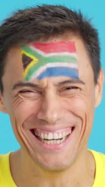 Stüdyoda, yüzünde kameraya gülümseyen Güney Afrika bayrağı olan beyaz bir adamın krom görüntüsü var.