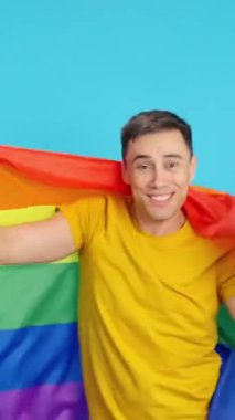 Lgbt gökkuşağı bayrağı sallayan, dans eden ve gülen bir adamın mavi arka planlı videosu stüdyoda. Çerçeve boş başlar, adam aşağıdan görünür ve altta kaybolur..