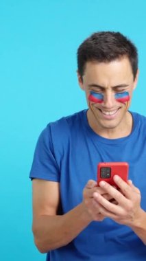 Yüzünde Haiti bayrağı olan Haitili bir destekçinin krom arkaplanlı stüdyo videosu, cep telefonuna bakıp gülümsüyor ve krom tuşlu ekranla kameraya gösteriyor.