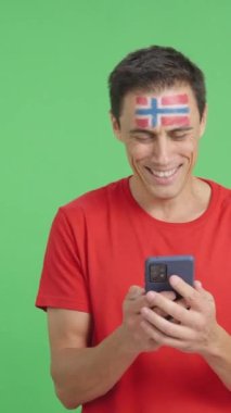 Norveçli bir destekçinin krom arkaplanlı stüdyo videosu. Yüzünde Norveç bayrağı var. Cep telefonuna bakıp gülümsüyor ve ekranı krom tuşlu gösteriyor.