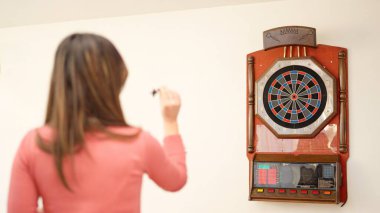 Rear view of a young caucasian woman playing darts at home clipart
