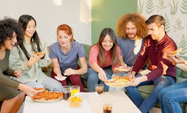 Çok ırklı arkadaşlar evde birlikte pizza yiyerek rahatlıyor ve gülümsüyorlar.