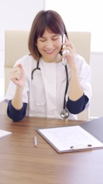 Kadın bir doktorun klinikteki bir masada oturan başarılı bir prosedürü kutlamak için telefonda konuştuğu yavaş çekim videosu.