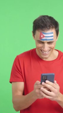 Yüzünde Küba bayrağı olan bir Kübalı destekçinin krom arkaplanlı stüdyo videosu, cep telefonuna bakıp gülümsüyor ve ekranda kroma tuşuyla onu gösteriyor.