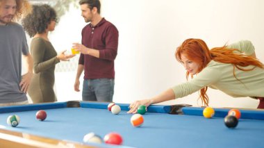 Kızıl saçlı kadın evde arkadaşlarıyla bilardo oynuyor.