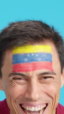 Stüdyoda, yüzünde Venezuela bayrağı olan ve kameraya gülümseyen bir adamın krom görüntüsü var.