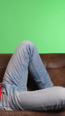 Genç adam yeşil ekran arka planına karşı akıllı telefon kullanarak kanepede yatıyor.