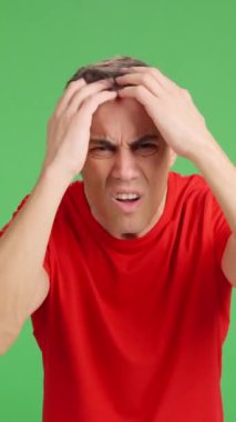 Kırmızı gömlekli kaygılı bir adam saç dökülmesi yaşıyor, yeşil ekranda kafasını kontrol ediyor, endişe ve hayal kırıklığını dile getiriyor.