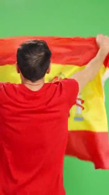 Sırtı kırmızı tişörtlü bir adam yeşil ekranda İspanyol bayrağı taşıyor.