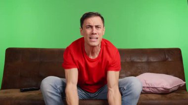 Evdeki kanepede oturan bir adam arkadaşlarıyla TV 'de maç izliyor. Ekibi bir şansı kaçırır ve umutsuzluğa kapılır. Orta atış, kırmızı gömlek ve yeşil krom anahtar arka plan..