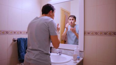 Nemlendirici yüz kremi süren bir adam banyo aynasında kendine bakıyor.
