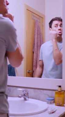 Adam dişlerini fırçalıyor ve banyo aynasında yüzünü yıkıyor.