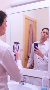 Narsist işadamı banyo aynasında akıllı telefonuyla fotoğraf çekiyor. Saçını ve kıyafetlerini düzeltiyor.