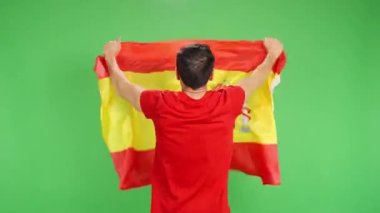 Sırtı kırmızı tişörtlü bir adam yeşil ekranda İspanyol bayrağı taşıyor.
