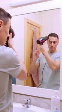 Adam banyodaki aynaya bakarken saç kurutma makinesi kullanıyor, özel bakım ve bakım rutinine odaklanıyor.