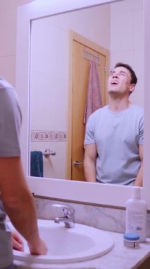 Kendine güveni olmayan bir adam banyodaki aynada saçını tarıyor ve hayal kırıklığına uğramış hissediyor.