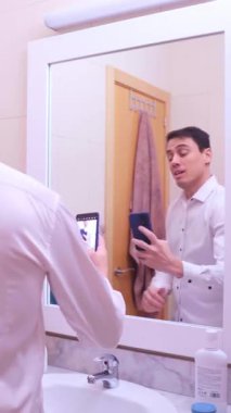 Kendine güveni olmayan işadamı banyo aynasına bakıyor ve akıllı telefonuyla selfie çekiyor.