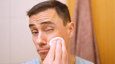 Genç adam yüz temizleme için pamuk ped kullanıyor, bir gözünü kırpıyor.