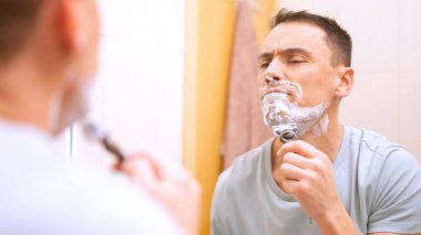 Sakalını tıraş köpüğü ve jiletle tıraş eden konsantre bir adam banyodaki aynaya bakıyor.
