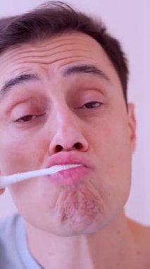 Yetişkin bir erkek dişlerini fırçalıyor. Banyodaki aynadaki yansımasına bakıyor.