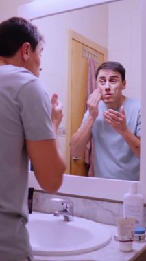 Nemlendirici yüz kremi süren bir adam banyo aynasında kendine bakıyor.