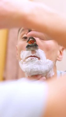 Beyaz bir adam, banyo aynasının önünde tıraş olurken bıyığını tıraş ediyor.