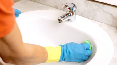 Eldiven giyen hademe yeşil süngerle beyaz lavaboyu temizliyor.