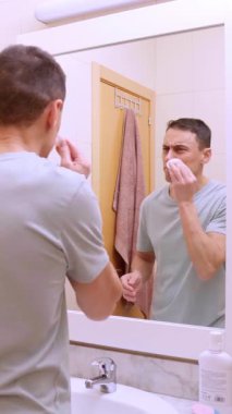 Adam, fayanslı bir banyoda pamuk pedleri ve yüz ürünleri kullanarak cilt temizleme rutinini gerçekleştiriyor.