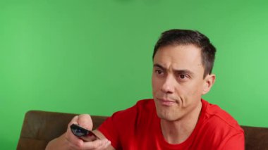 Kanepede elinde uzaktan kumandayla oturan, sinirli ifadeyle kanalları çeviren, yeşil ekran arka planında elektrik veren bir adam.