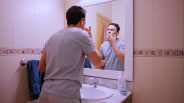 Genç adam banyo aynasının önünde kakao yağıyla dudaklarını nemlendiriyor.