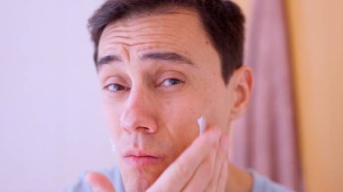 Genç adam rutin cilt bakımı için yüzüne nemlendirici krem sürüyor.