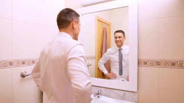 İş adamı işe hazırlanıyor, banyo aynasında yansımasına gülümsüyor, kendinden emin ve başarılı hissediyor.