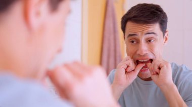 Diş temizliği için diş ipi kullanan bir adam. Banyo aynasının önünde ağız hijyenini koruyor.